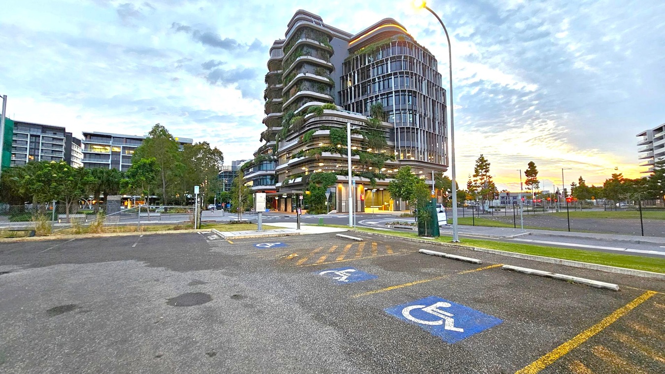 A modern multi-storey building with curved architecture and greenery on its balconies is in the centre. In the foreground, there is a parking area with marked disabled parking spaces. To the left, there are more buildings and trees, while to the right, a street with a lamppost and additional trees. The background shows a partly cloudy sky with a hint of sunset colours.