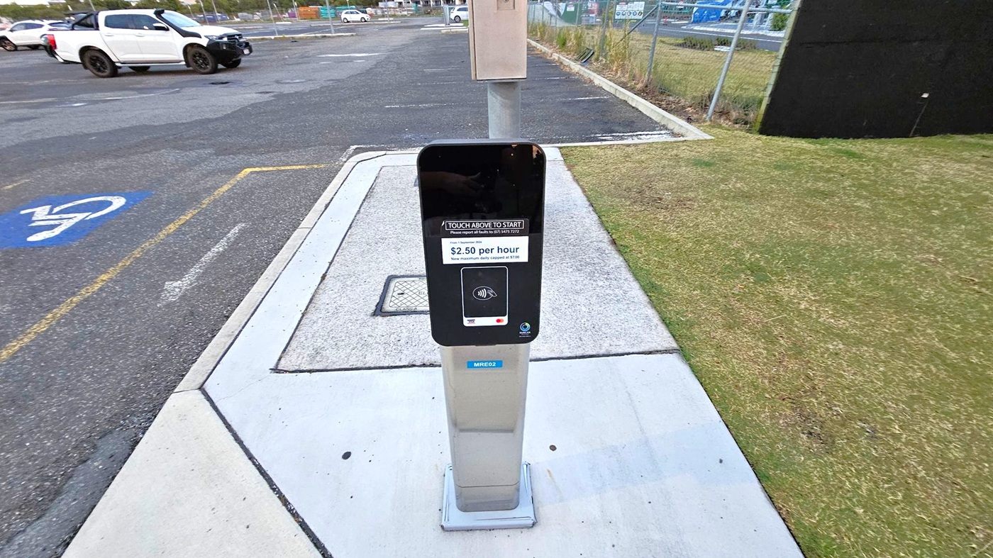 A parking payment machine is in the centre, mounted on a grey post with a touch interface displaying a rate of $2.50 per hour. To the left, a white pickup truck is parked in a marked bay on an asphalt surface, with a disabled parking symbol visible. On the right, there is a patch of grass with some brown areas. In the background, a fence and more parked vehicles are visible. The ground is a mix of concrete and asphalt.