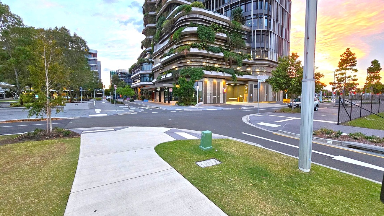 A modern urban scene with a curved building on the right, featuring multiple levels adorned with greenery. The foreground has a concrete path and a small grassy area with a green utility box. A street with marked lanes and a pole is visible. On the left, there are more buildings and trees lining the street. The background shows a sunset with orange and pink hues.