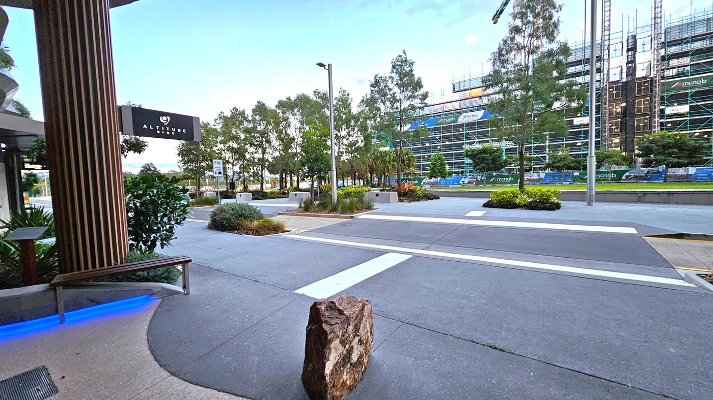 On the left, a building with a sign reading "Altitude" and a wooden column. A bench with blue lighting underneath is nearby. In the centre, a wide pavement with a large rock and a pedestrian crossing. Trees and shrubs line the area. On the right, a construction site with scaffolding and a building under development. The background features more trees and a clear sky.