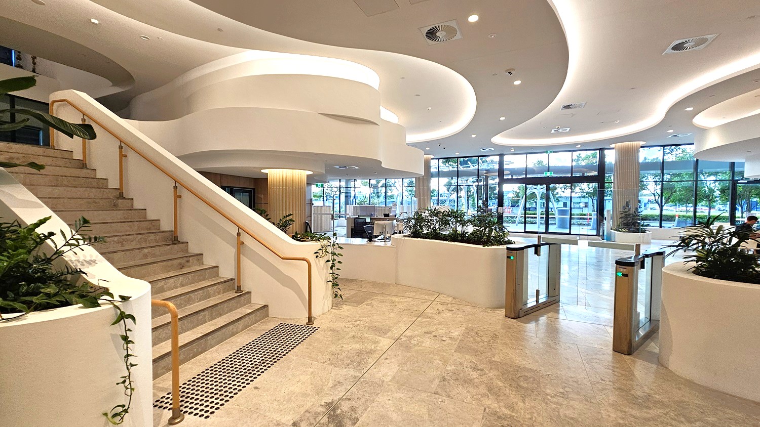 Spacious modern lobby with a curved staircase on the left, featuring a wooden handrail and stone steps. The floor is polished stone, and the ceiling has flowing, wave-like designs with recessed lighting. In the centre, there are planters with greenery and a security gate. Large glass windows in the background allow natural light to illuminate the space, with views of trees and a street outside.