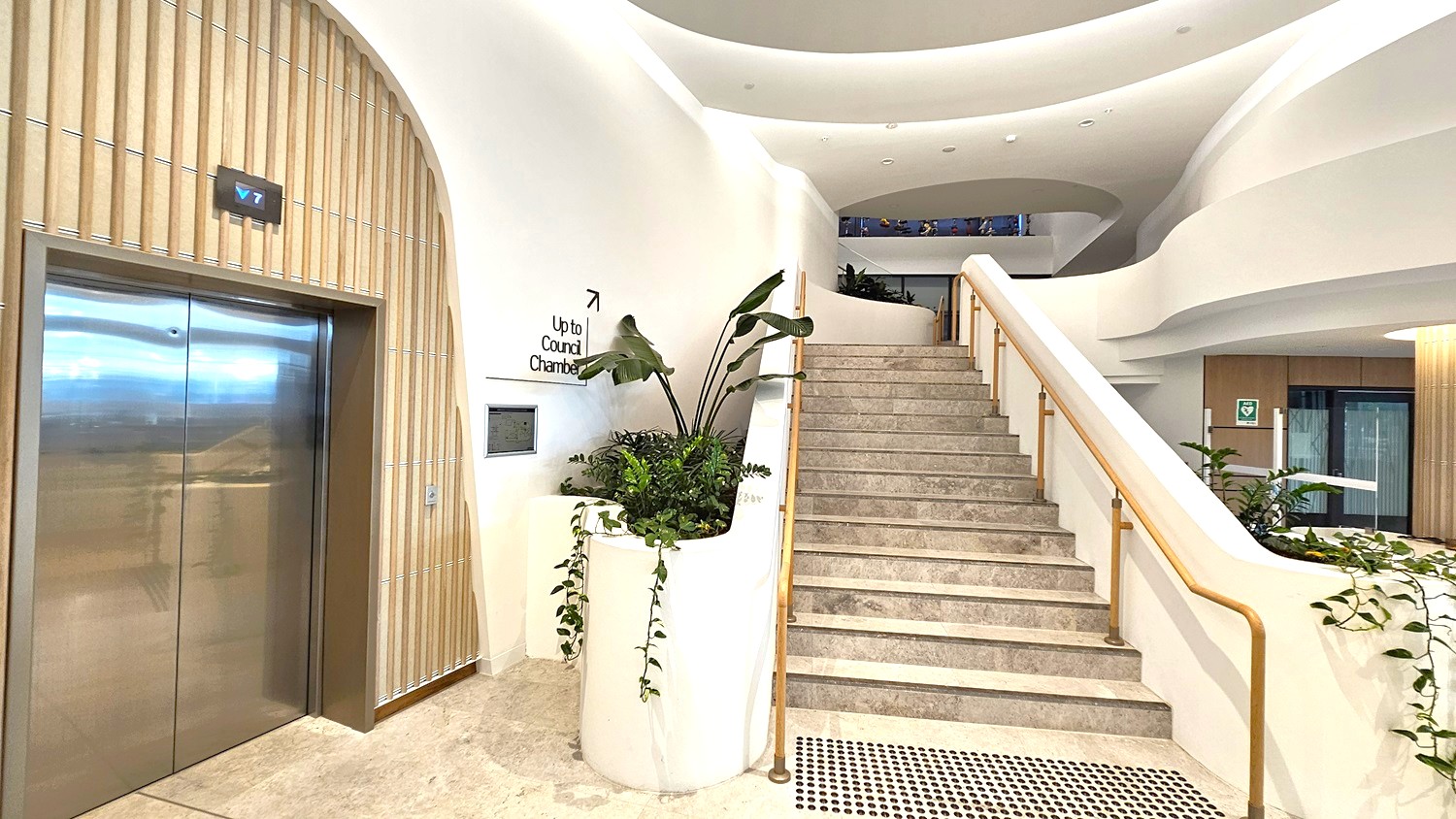 A modern interior with a staircase on the right, featuring a light stone finish and a sleek handrail. To the left, a stainless steel lift with a digital floor indicator above. Between the lift and the stairs, a planter with lush green foliage. The walls are smooth and white, with a sign indicating the direction to the council chamber. The ceiling has a curved, layered design, and the floor is a light stone with a textured mat near the stairs.
