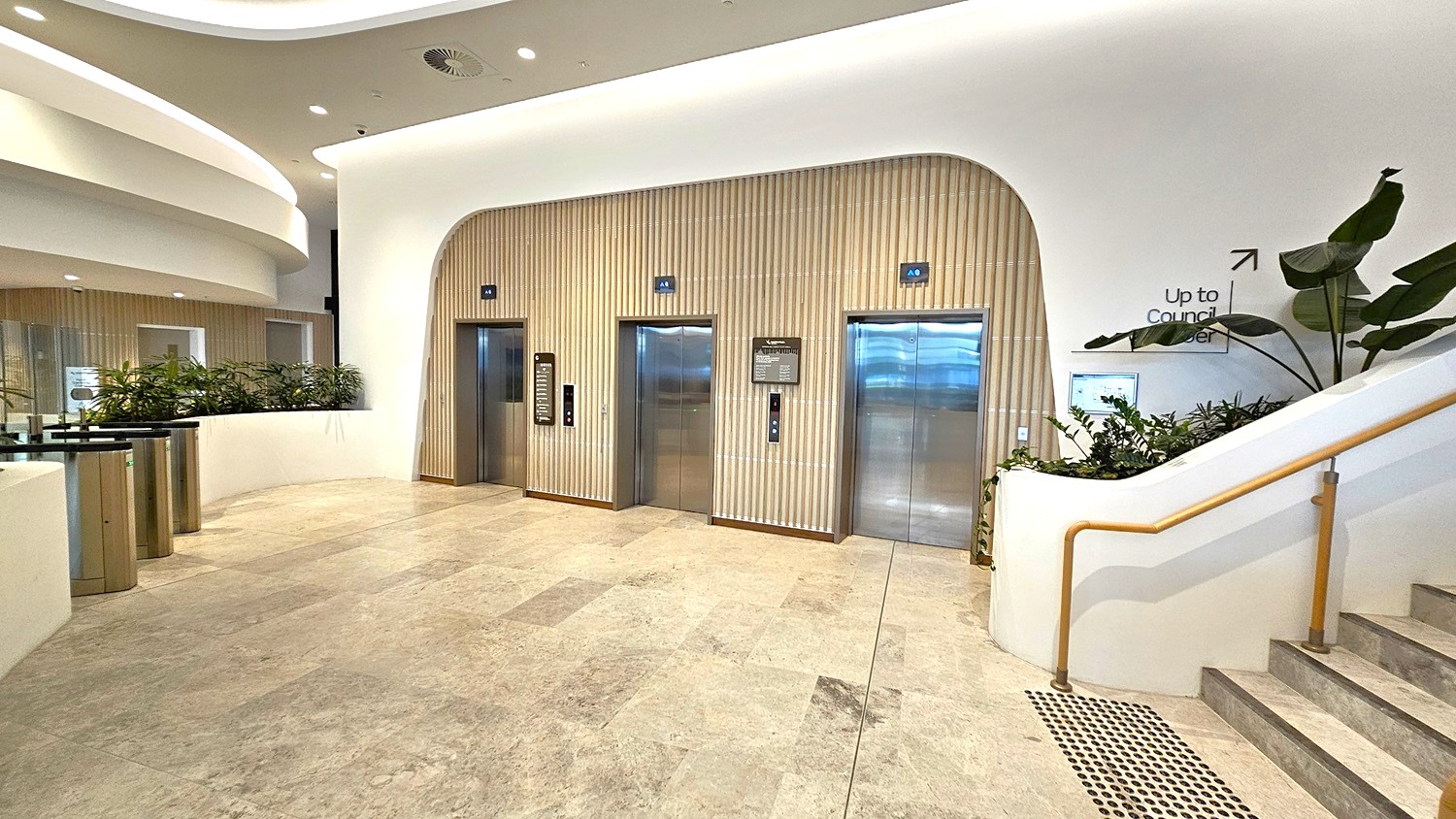 A modern interior with three lifts in the centre, set against a wall with vertical wooden panelling. To the right, a staircase with a wooden handrail leads upwards, accompanied by a sign indicating "Up to Council Level". There are potted plants along the staircase and near the lifts. On the left, a curved wall with more plants and a sleek turnstile. The floor is made of light-coloured stone tiles, and the ceiling features recessed lighting with a smooth, curved design.