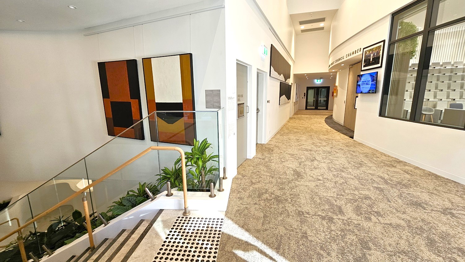 A modern interior with a staircase on the left featuring a glass railing and wooden handrail, surrounded by potted plants. The wall displays abstract artwork. On the right, a carpeted hallway leads to a distant doorway, with framed pictures and a digital screen mounted on the wall. A large window reveals a room with tiered seating. The ceiling is smooth and white, with recessed lighting.