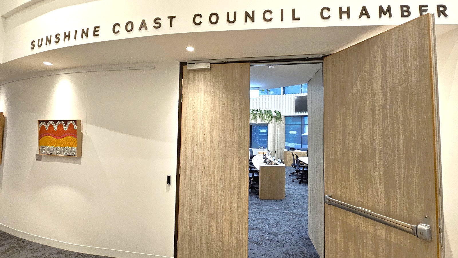 A hallway with a curved white wall on the left, featuring a colourful abstract painting. Above, the words "SUNSHINE COAST COUNCIL CHAMBER" are displayed. A wooden door is partially open, leading into a room with a large, curved conference table surrounded by chairs. The room has blue carpet and large windows with vertical blinds. Green plants are visible on a shelf near the windows.