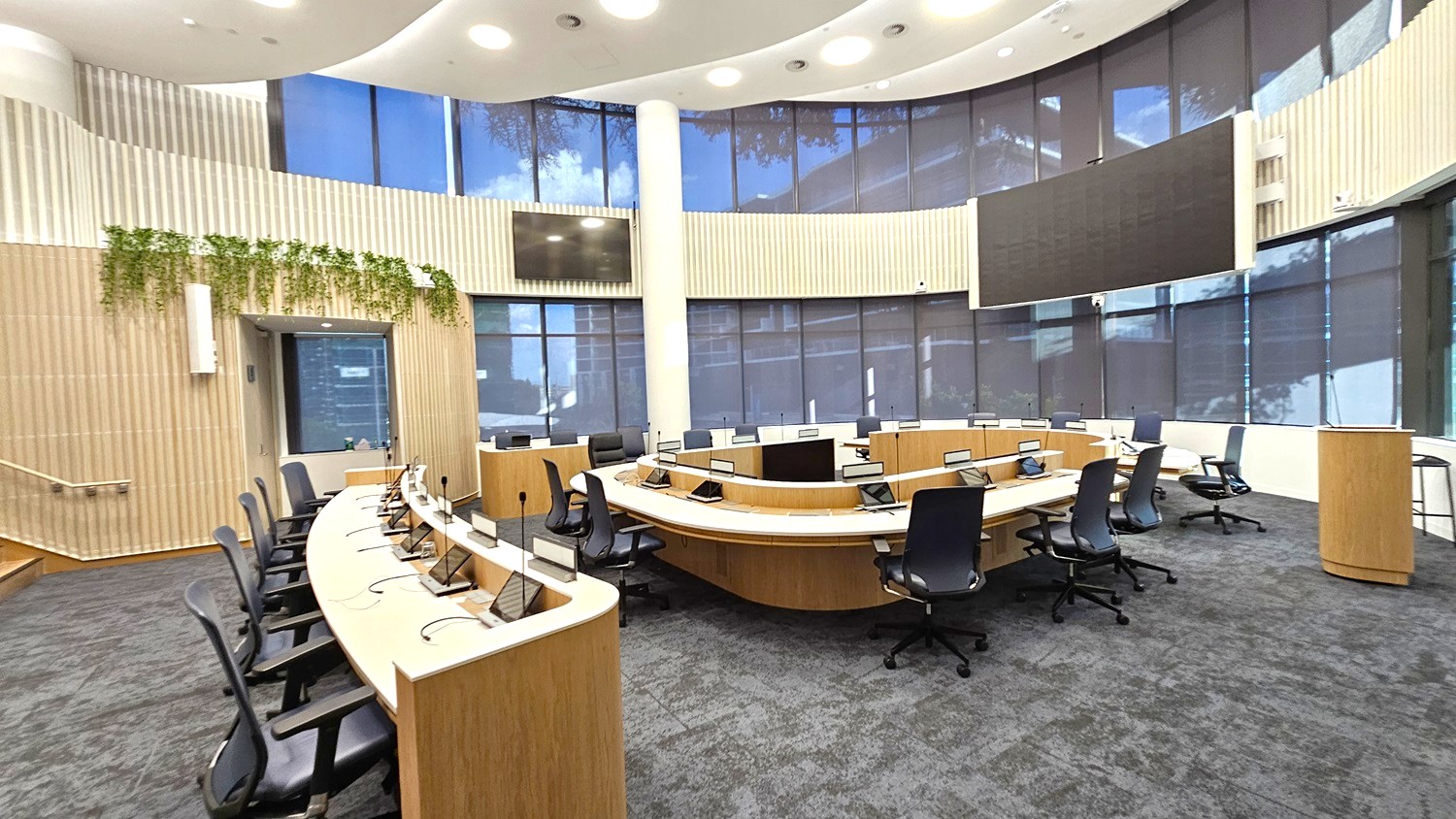 A modern conference room with a circular arrangement of wooden desks and black office chairs. In the centre, there are multiple computer monitors and microphones. The walls are lined with large windows covered by blinds, and a strip of greenery decorates the left wall. The ceiling is white with recessed lighting. The floor is carpeted in a dark grey tone. A large screen is mounted on the right wall, and a wooden podium stands nearby.