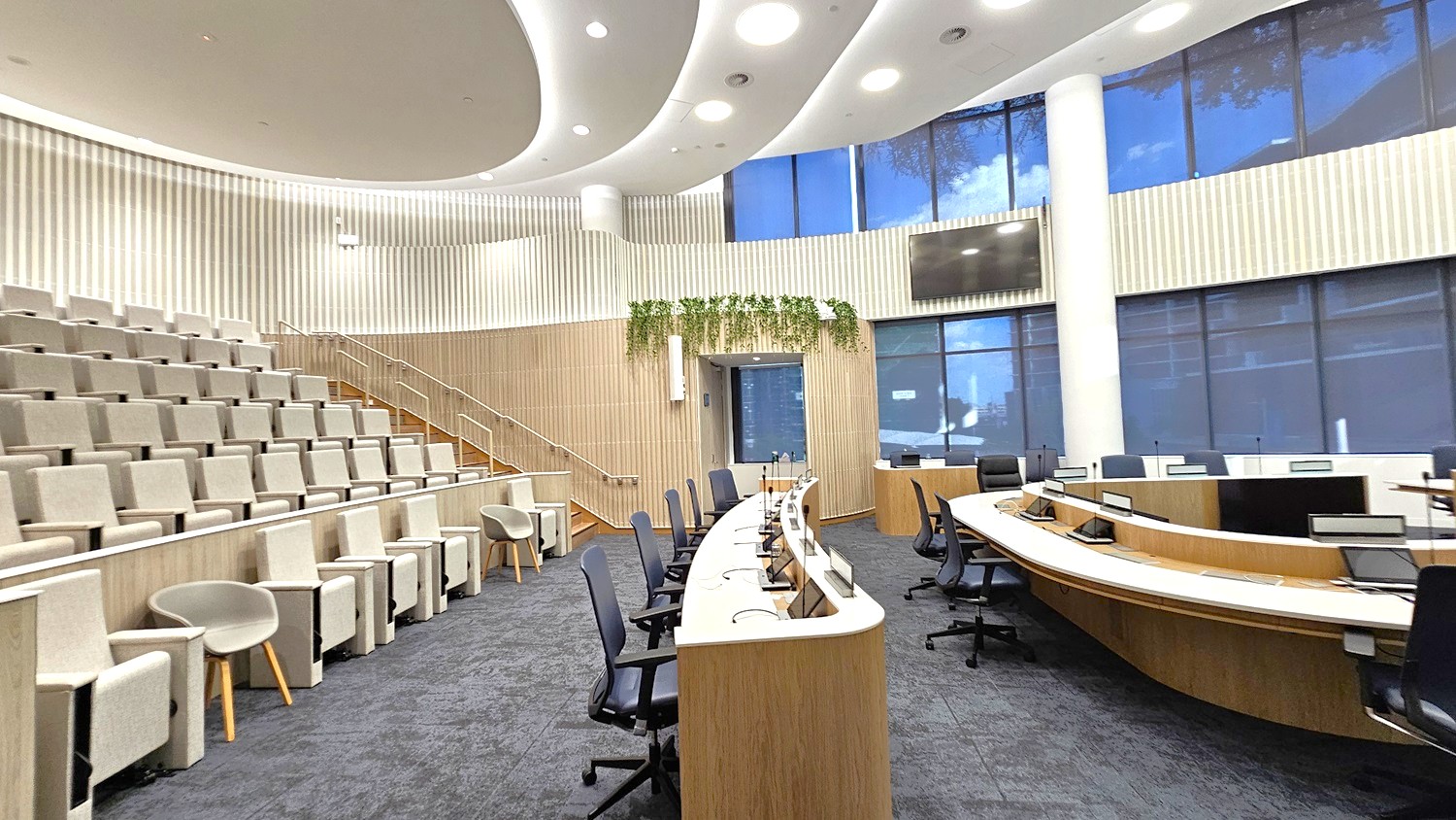 A modern conference room with tiered seating on the left, featuring light grey upholstered chairs. In the centre, there are curved wooden desks with black office chairs and computer monitors. The back wall has vertical wooden panelling and a large screen, with plants hanging above a doorway. The right side has large windows with blue-tinted glass. The ceiling is white with recessed lighting, and the floor is covered in dark grey carpet.