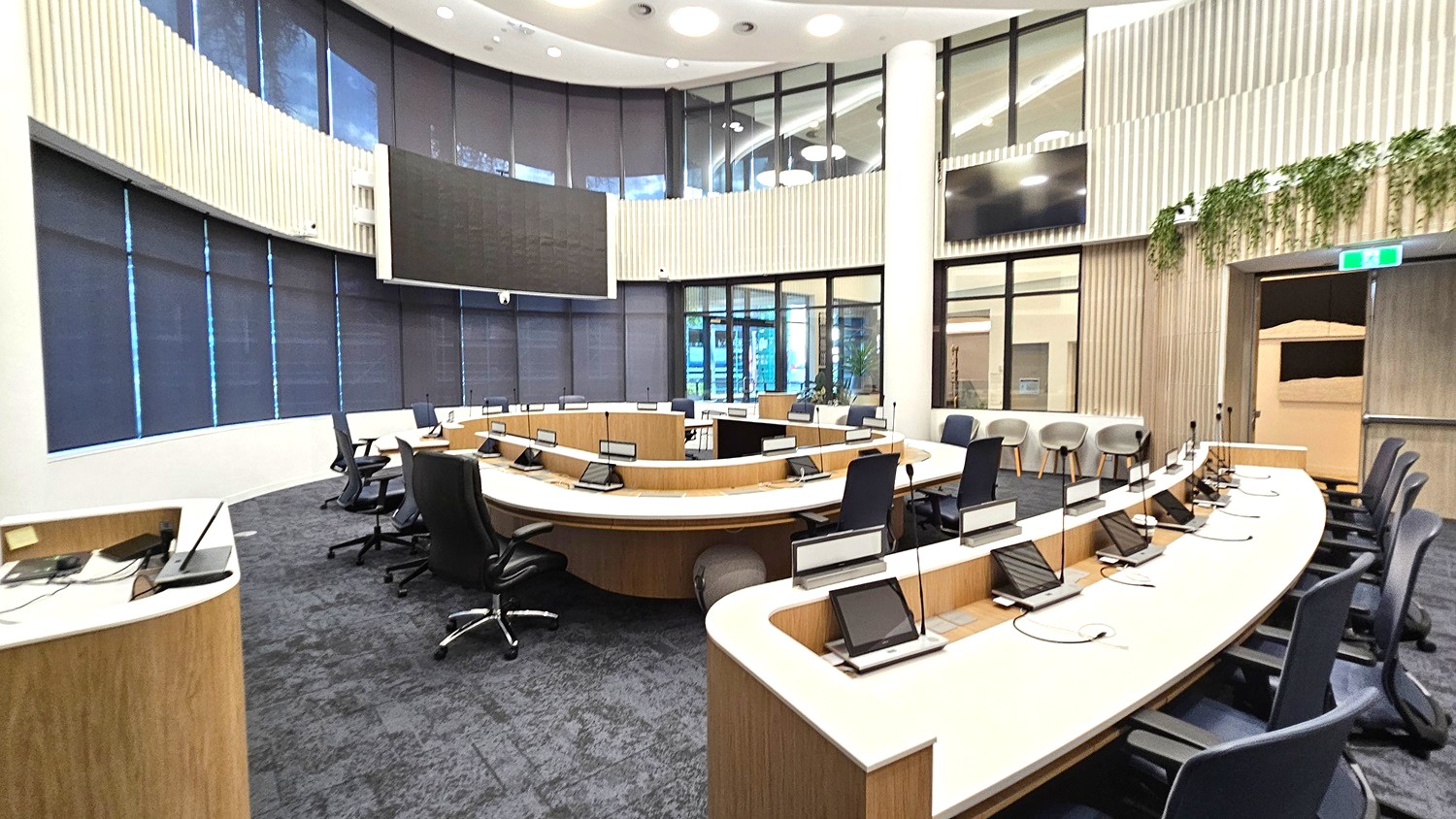 A modern conference room with a semi-circular arrangement of desks equipped with microphones and screens. Black office chairs are positioned around the desks. The floor is carpeted in a dark grey tone. Large windows with dark blinds line the curved wall in the background. The ceiling is high with circular lights. On the right, there are additional chairs and a wall with vertical wooden panelling and some greenery. A large screen is mounted on the wall.