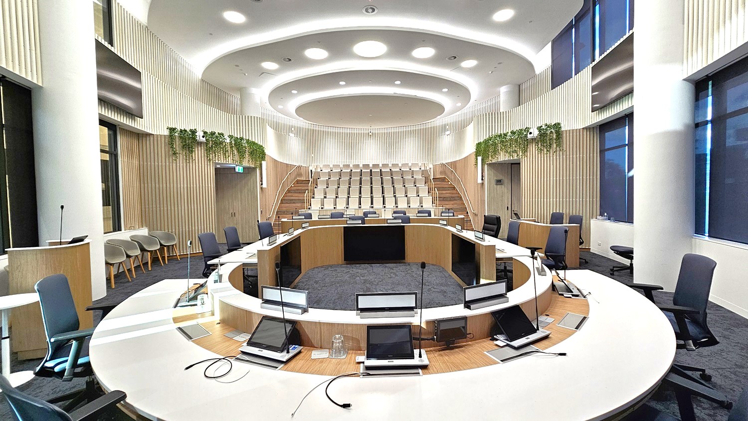 A modern conference room with a circular seating arrangement. In the foreground, there are several chairs and desks equipped with microphones and screens. The middle features a central circular area with additional seating. The background shows tiered seating with light-coloured chairs. The walls are adorned with vertical wooden panels and greenery. The ceiling has circular light fixtures, and the floor is carpeted in a dark shade.