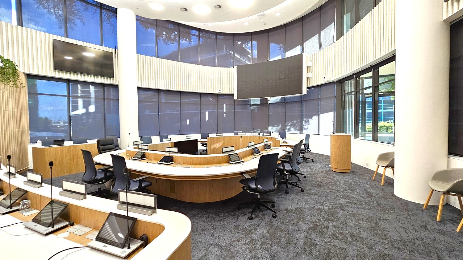 A modern conference room with a semi-circular arrangement of desks and black office chairs. Each desk has a microphone and a small screen. Large windows with dark blinds line the curved wall, and a large screen is mounted above. The floor is carpeted in dark grey, and the ceiling is white with circular lights. A wooden podium stands near the windows, and two beige chairs are positioned against the wall.