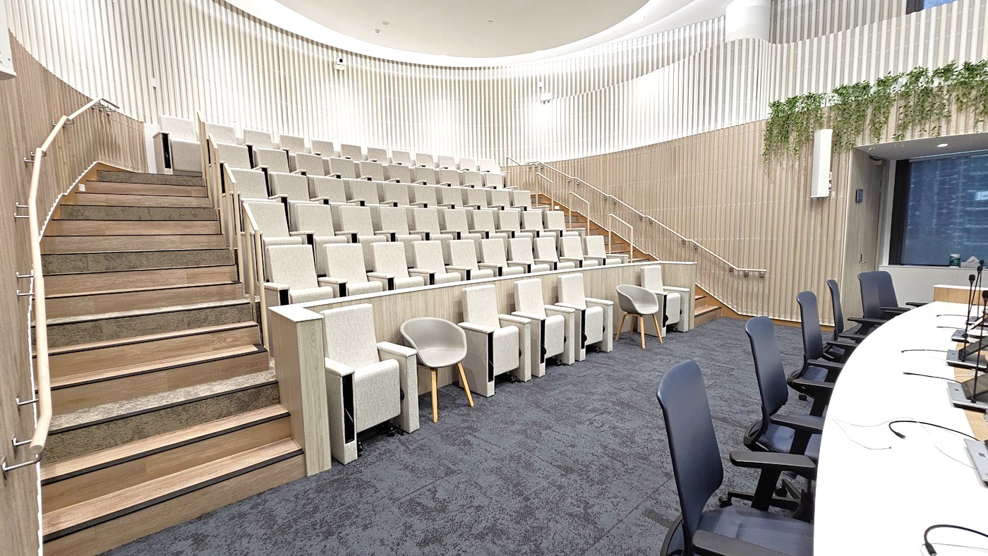 A modern lecture hall with tiered seating on the left, featuring light green chairs and wooden steps with handrails. On the right, a curved table with black chairs and microphones. The walls are lined with vertical wooden panels, and there are plants hanging on the right side. The floor is carpeted in dark grey, and the ceiling is light with recessed lighting.