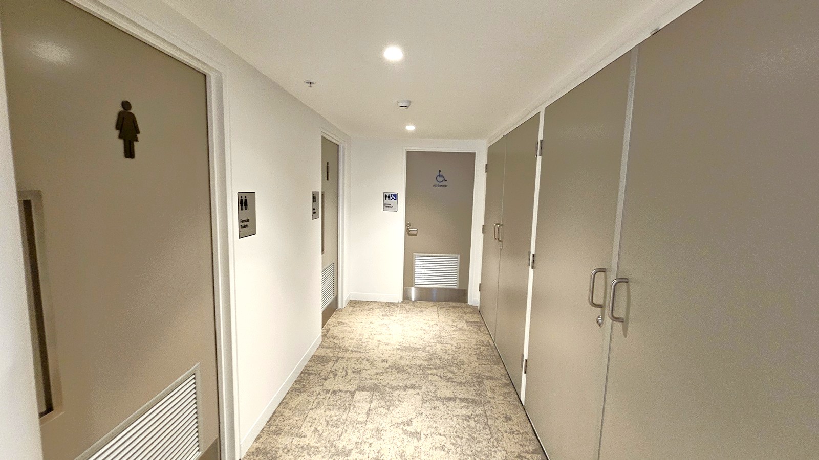 A corridor with beige walls and a light-coloured ceiling featuring recessed lighting. On the left, there are doors with gender symbols, and a sign indicating a women's restroom. On the right, there are more doors with handles. At the end of the corridor, a door with a wheelchair symbol and a sign for an accessible restroom is visible. The floor is covered with a patterned carpet.