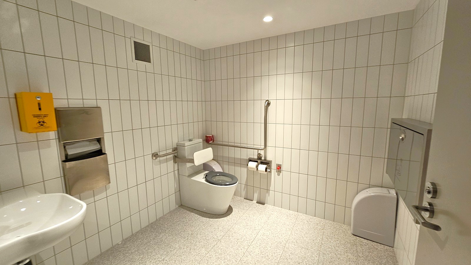 A public restroom with white tiled walls and a speckled grey floor. In the centre, a toilet with a green seat is equipped with support rails. To the right, a sanitary disposal bin is mounted on the wall. On the left, a sink is partially visible, along with a stainless steel paper towel dispenser and a yellow biohazard disposal box. The ceiling is plain with recessed lighting.