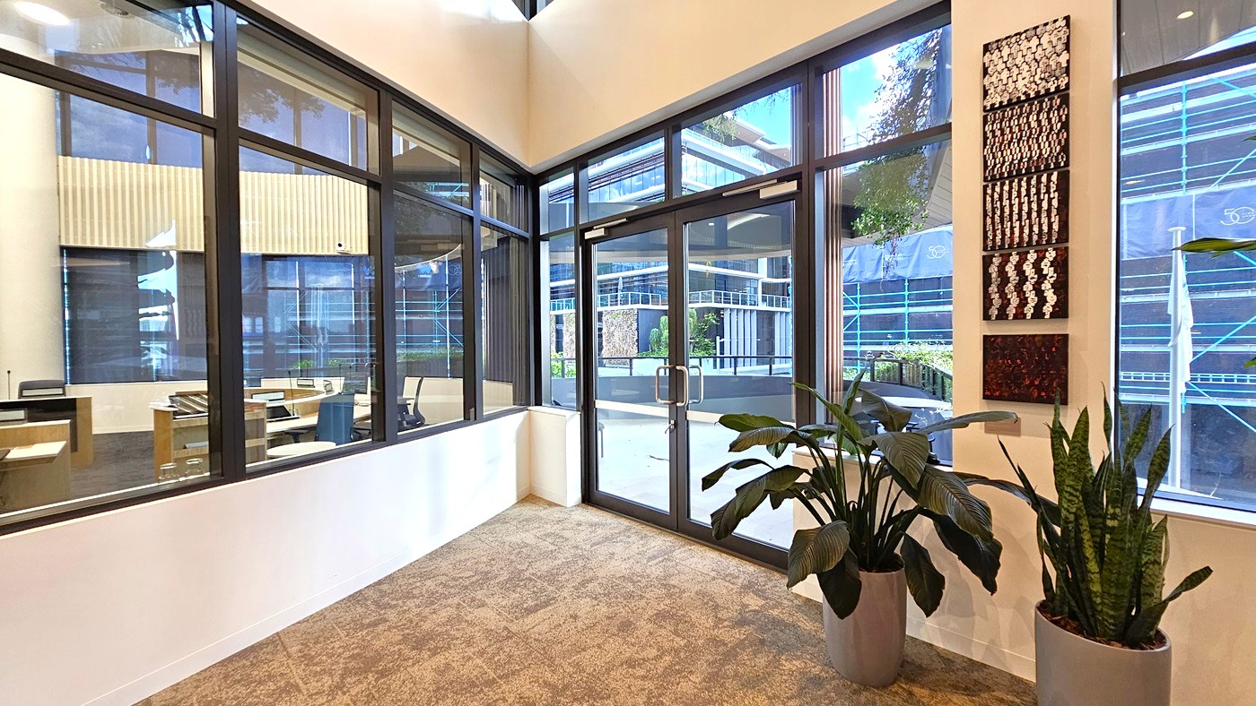 A modern office interior with large glass windows and doors. On the left, an office area with desks and chairs is visible through the glass. The floor is carpeted in a neutral tone. On the right, two large potted plants are placed near the windows, and a series of abstract artworks are mounted on the wall. Outside, a view of a contemporary building with glass and metal elements can be seen. The ceiling is not visible.