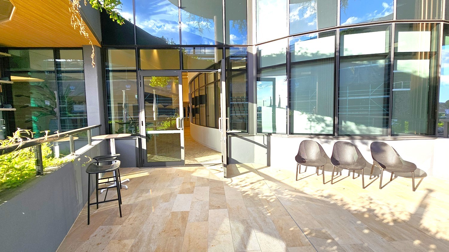 On the left, there is a small round table with a black stool next to a glass railing with plants. The middle features a glass door entrance with reflections of the surroundings. On the right, three black chairs are lined up against a wall with large windows. The floor is made of light-coloured tiles, and the ceiling is partially visible with a wooden finish. Shadows of trees are cast on the ground and wall.