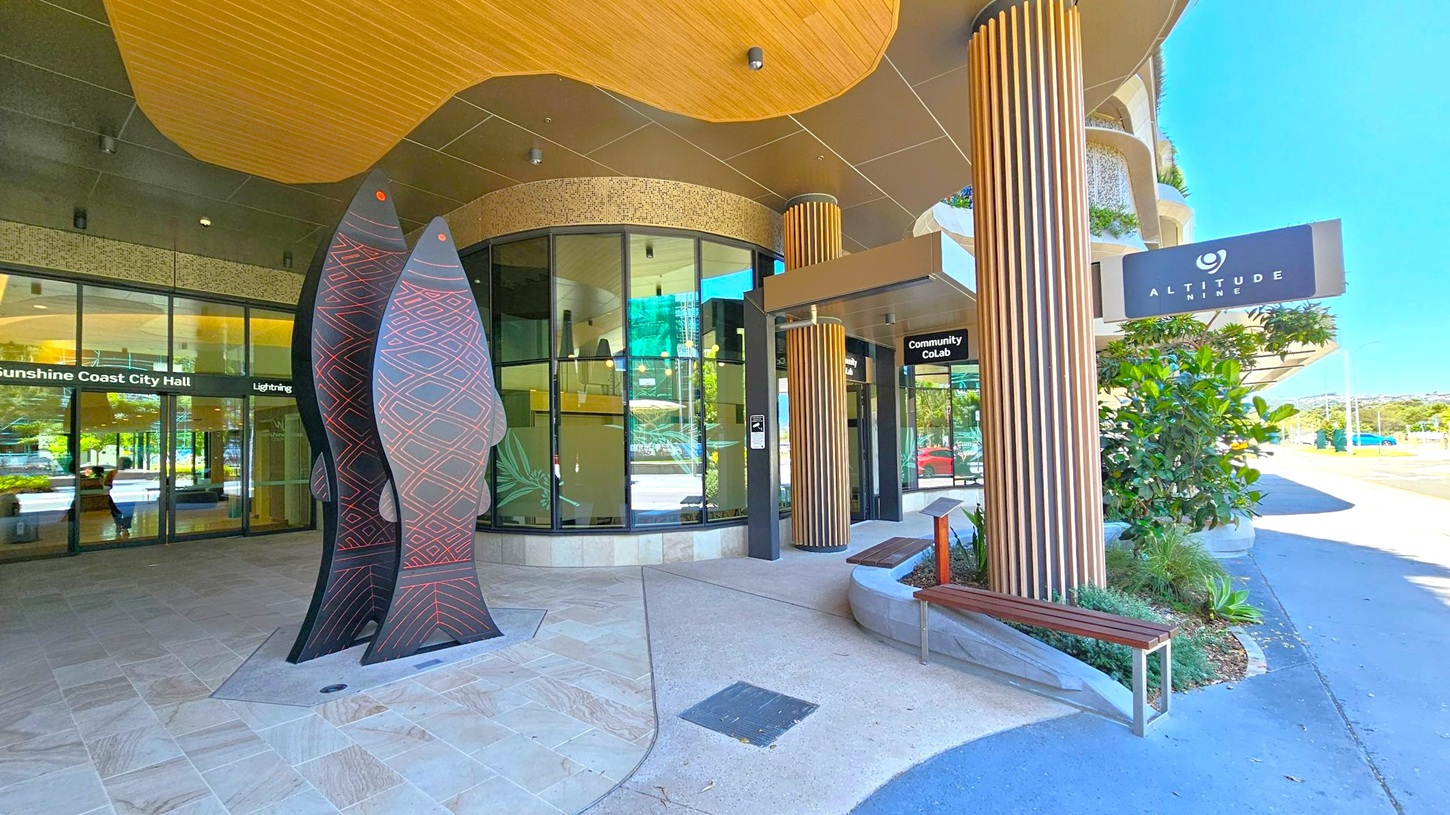Entrance area with a modern design. On the left, there are large glass doors with signage for "Sunshine Coast City Hall" and "Lightning." In the centre, a tall, artistic sculpture resembling fish with geometric patterns stands prominently. On the right, a sign for "Altitude Nine" is visible above a wooden bench and potted plants. The ceiling features a wooden panel design, and the floor is a mix of stone and concrete. The background shows a street and distant landscape.