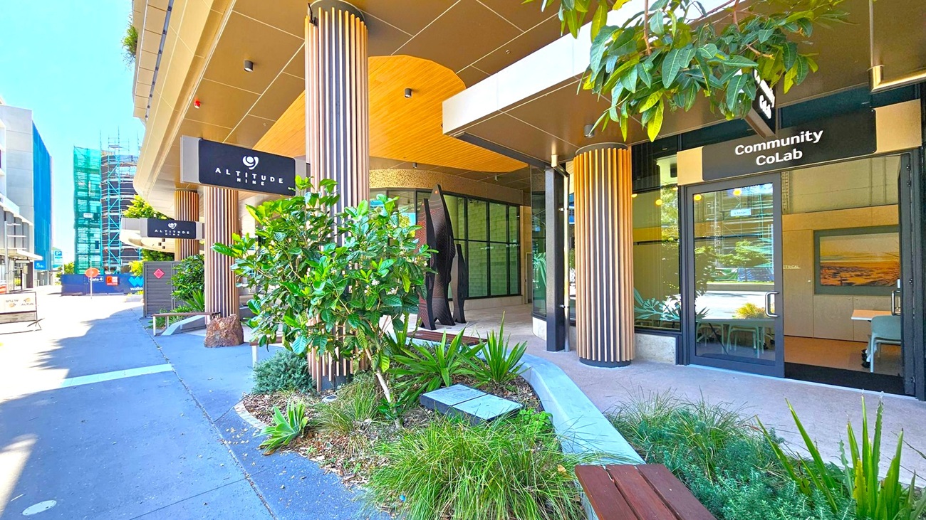 Entrance to a modern building with a sign reading "Altitude Nine" on the left and "Community CoLab" on the right. The facade features vertical wooden slats and large glass windows. A small garden with various green plants and a bench is in the foreground. The pavement is grey, and there are construction barriers and scaffolding visible in the background on the left. The ceiling is smooth and light-coloured, complementing the building's contemporary design.