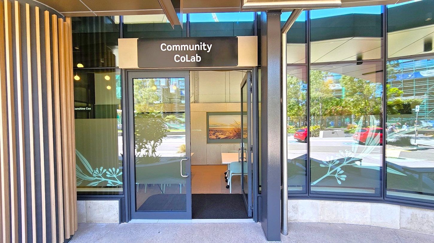 Entrance to a building with a sign reading "Community CoLab" above the door. The door is glass with metal handles, and there are large windows on either side reflecting trees and parked cars outside. Inside, a framed picture is visible on the wall, and there are tables and chairs. The exterior features vertical wooden slats on the left and a concrete floor.