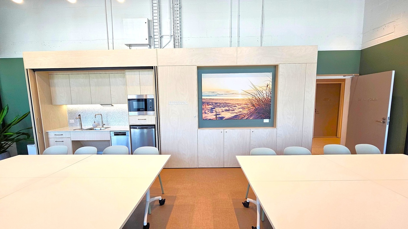 A modern office space with a kitchenette on the left, featuring a sink, microwave, and dishwasher. Above the kitchenette are white cabinets. In the centre, a large framed photograph of a beach scene is displayed. To the right, an open door leads to another room. In the foreground, there are white tables with chairs on wheels. The floor is carpeted, and the walls are painted in a mix of white and green. Ceiling lights are visible above.