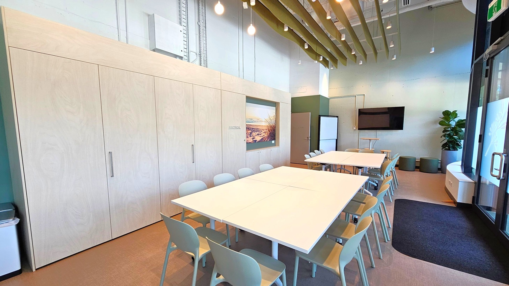 A modern meeting room with light wood cabinets on the left wall, featuring a landscape picture. In the centre, there are two large white tables surrounded by light grey chairs. The right side has a wall-mounted TV, a whiteboard, and a potted plant. The ceiling has exposed beams and hanging lights. The floor is carpeted, and there is a glass door on the right with a dark mat in front of it.