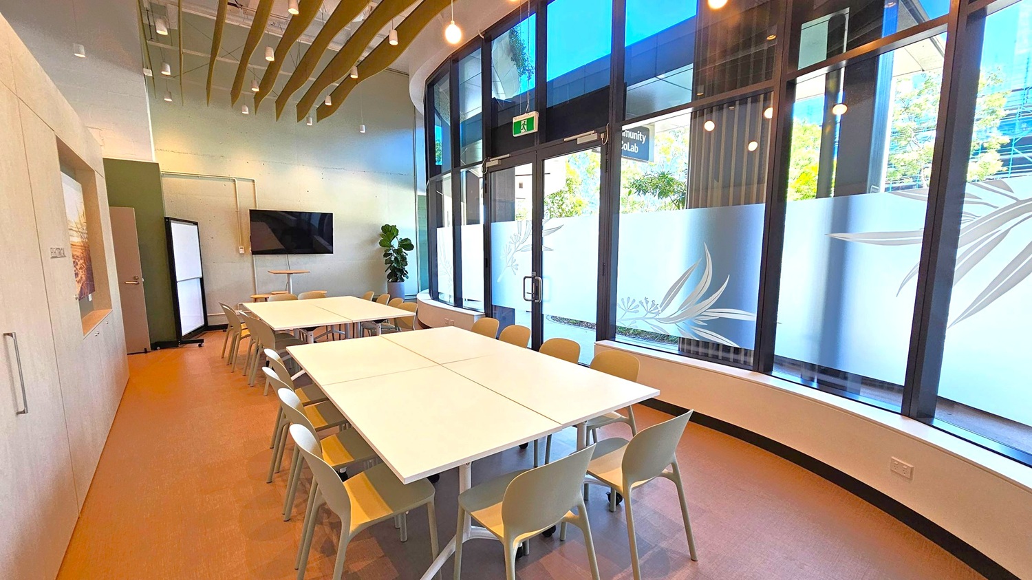 A modern meeting room with a large rectangular table in the centre surrounded by beige chairs. On the left, there is a wall with a door and a mounted flat-screen TV. The right side features large windows with frosted designs, allowing natural light to enter. A potted plant is near the corner. The ceiling has wooden slats and hanging lights, and the floor is carpeted in a light brown tone.