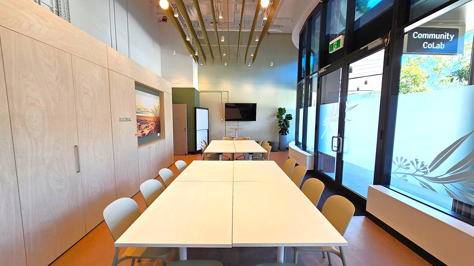 A modern meeting room with a long table and chairs in the centre. On the left, there are light wooden cabinets with a landscape picture. The right side features large windows with frosted designs and a "Community CoLab" sign. A TV is mounted on the back wall, and a potted plant is nearby. The ceiling has exposed beams and hanging lights. The floor is wooden, and the room has a clean, minimalist design.