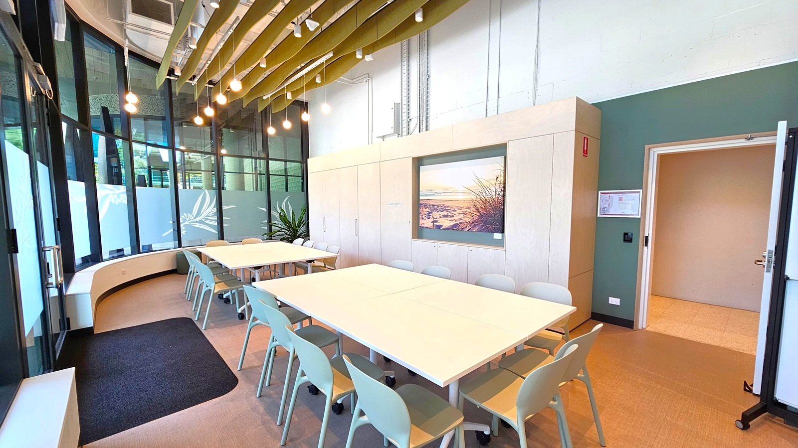 A modern meeting room with a large rectangular table in the centre surrounded by light grey chairs. On the left, there are tall windows with frosted glass panels featuring leaf patterns. The ceiling has exposed beams and hanging lights. On the right, a door is open, leading to another room. A large photograph of a beach scene is mounted on the wall. A plant is placed near the windows, and the floor is carpeted in a neutral tone.