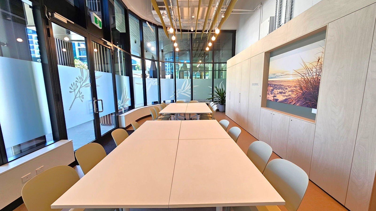 A modern meeting room with large glass windows on the left, featuring frosted designs and an exit sign above the door. In the centre, there are two rectangular tables with beige chairs around them. The right wall has light wood panelling with a large framed picture of a beach scene. A potted plant is placed near the windows. The ceiling has exposed pipes and hanging lights, and the floor is a light brown colour.