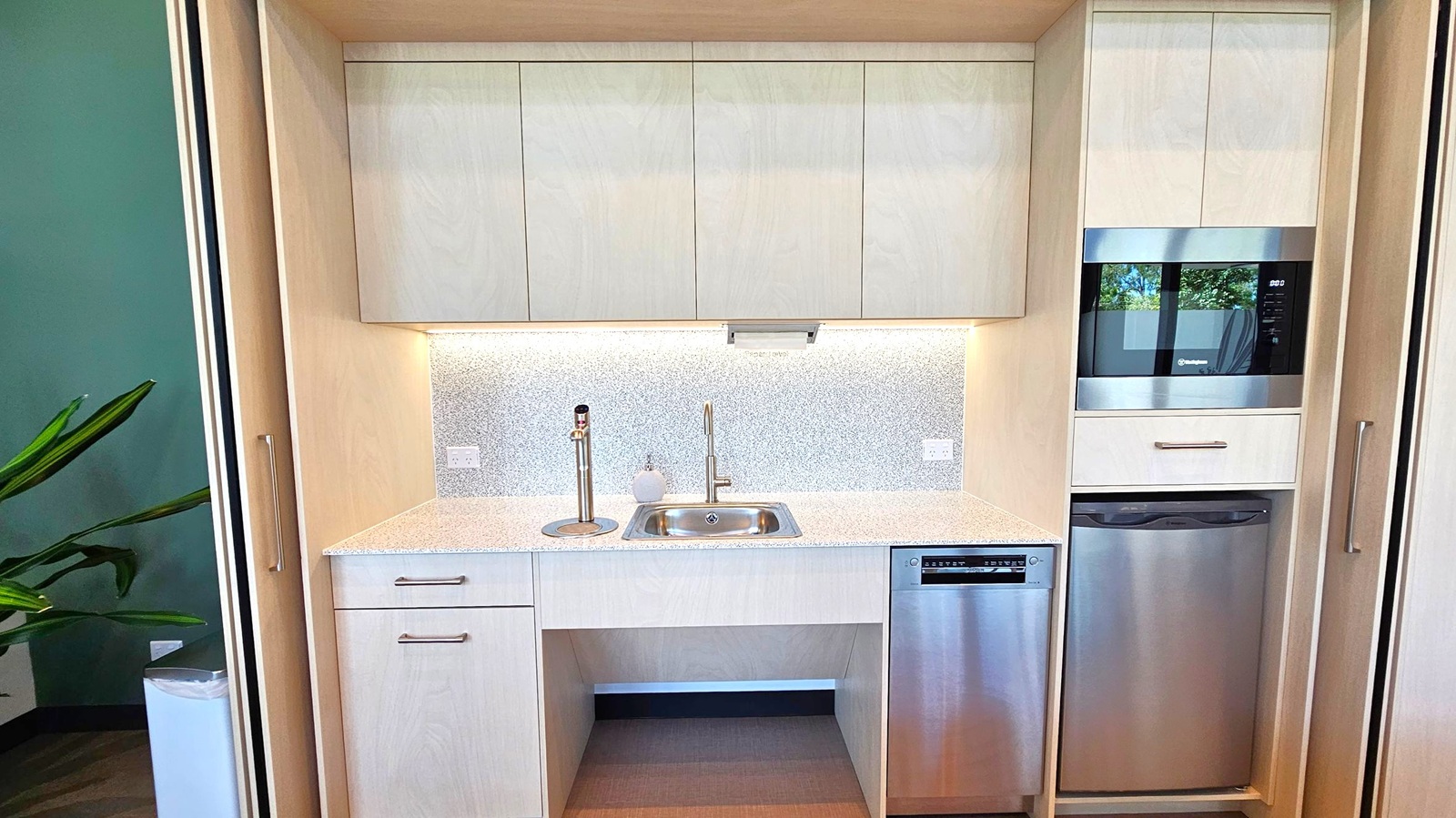A compact kitchen area with light wood cabinetry. In the centre, a small stainless steel sink with a modern tap is set into a speckled countertop. Above, there are white cabinets with a subtle sheen. To the right, a built-in microwave and a dishwasher below it, both with stainless steel finishes. On the left, a green wall with a large plant partially visible. The floor is a light wood, and the ceiling is not visible.