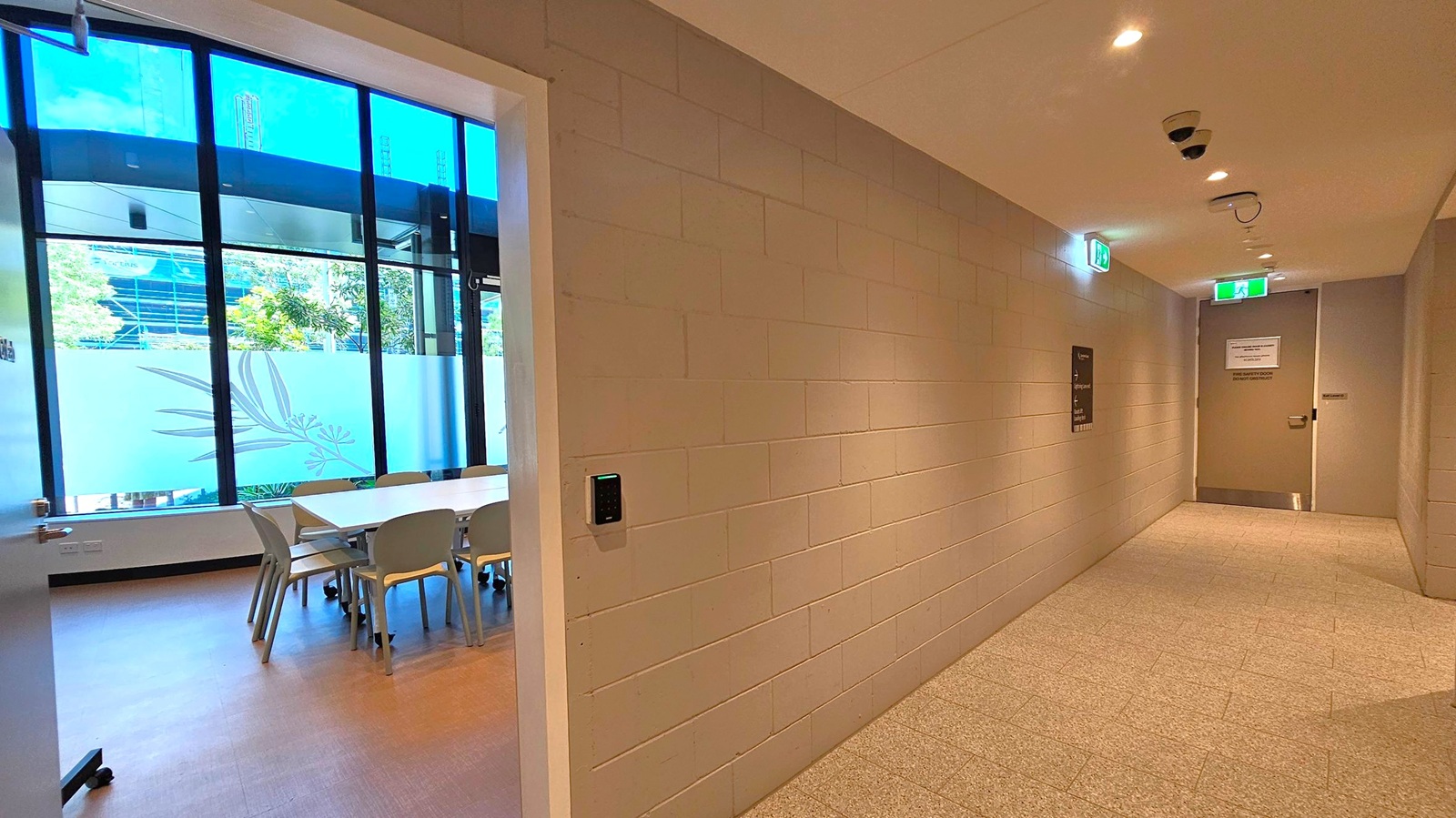 On the left, a room with large windows features a long table surrounded by chairs. The windows have frosted designs and offer a view of trees and a building outside. On the right, a corridor with beige brick walls leads to a door at the end. The ceiling has recessed lighting and security cameras. The floor is tiled in a light grey pattern. Exit signs are visible above the doors.