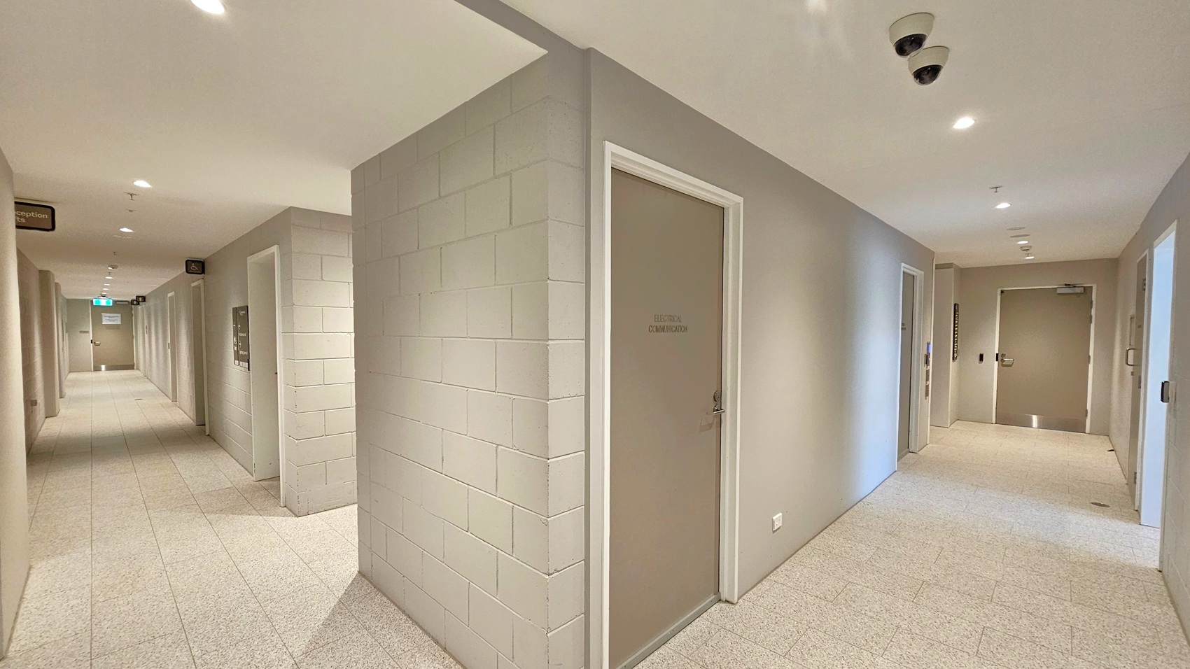 A corridor with light grey tiled flooring and a white ceiling featuring recessed lights and security cameras. On the right, a door labelled "Electrical Communication" is set into a light grey wall. The corridor extends to the left, with multiple doors and signs along the walls. At the far end, an exit sign is visible above a door. The walls are a mix of smooth and block textures, creating a clean and organised appearance.