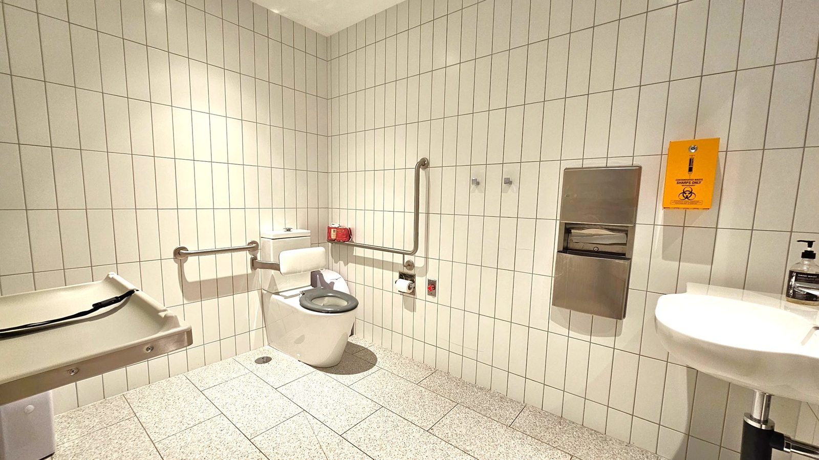 A public restroom with white tiled walls and a speckled grey floor. On the left, a baby changing station is mounted on the wall. In the centre, a toilet with support rails and a green seat cover is visible. Above the toilet, a red box and a toilet roll holder are attached to the wall. On the right, a stainless steel dispenser and a yellow sharps disposal bin are mounted. A white sink with a soap dispenser is also on the right. The ceiling is plain white.