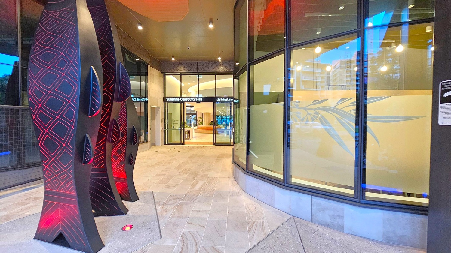 On the left, there are tall, black sculptures with red geometric patterns and small cut-out shapes. The floor is tiled with light-coloured stone. In the centre, glass doors lead to the Sunshine Coast City Hall, with a sign above reading "Lightning Lane". The interior is visible with a circular seating area and plants. On the right, large windows with frosted glass feature a leaf design. The ceiling is dark with recessed lighting.