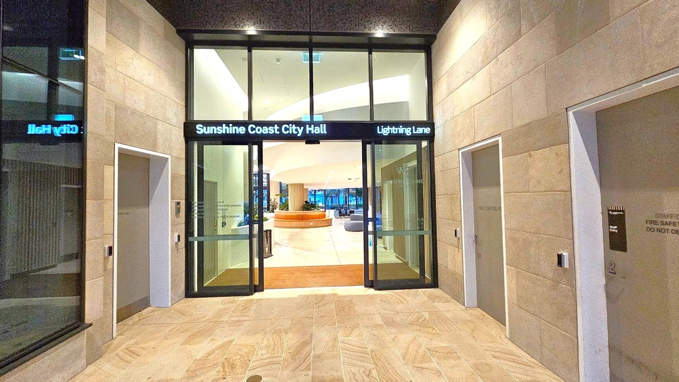 Entrance to a modern building with large glass doors labelled "Sunshine Coast City Hall" and "Lightning Lane". The walls are clad in light-coloured stone tiles. On either side of the entrance are doors marked "Fire Control Rm" and "Staff Entry". Inside, a spacious lobby with a circular seating area and plants is visible. The floor is tiled, and the ceiling features recessed lighting.