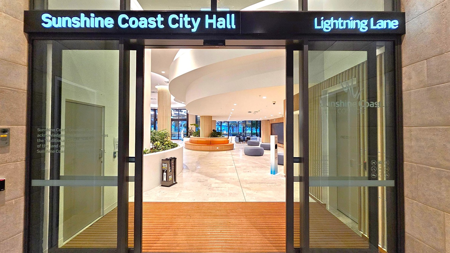 Entrance to Sunshine Coast City Hall with "Lightning Lane" signage above. Glass doors open to a spacious interior featuring a circular seating area with plants. The floor is light-coloured and polished, and the ceiling has a modern, curved design with recessed lighting. To the left, a wall-mounted text acknowledges traditional custodians. On the right, a digital display is visible. The background shows large windows with a view of trees outside.