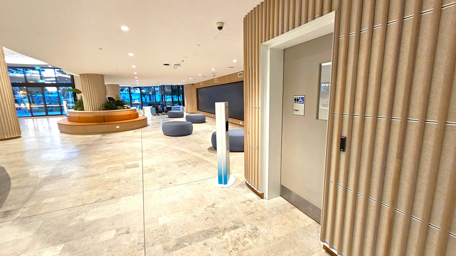 A modern indoor space with a light-coloured tiled floor and a white ceiling with recessed lighting. On the right, there's a door with a sign indicating accessible toilets. The wall features vertical wooden slats. In the middle, a tall, slim, gradient-coloured stand is visible. The left side has a circular seating area with plants, and further back, there are round, grey seating poufs. Large glass windows in the background reveal an outdoor view.
