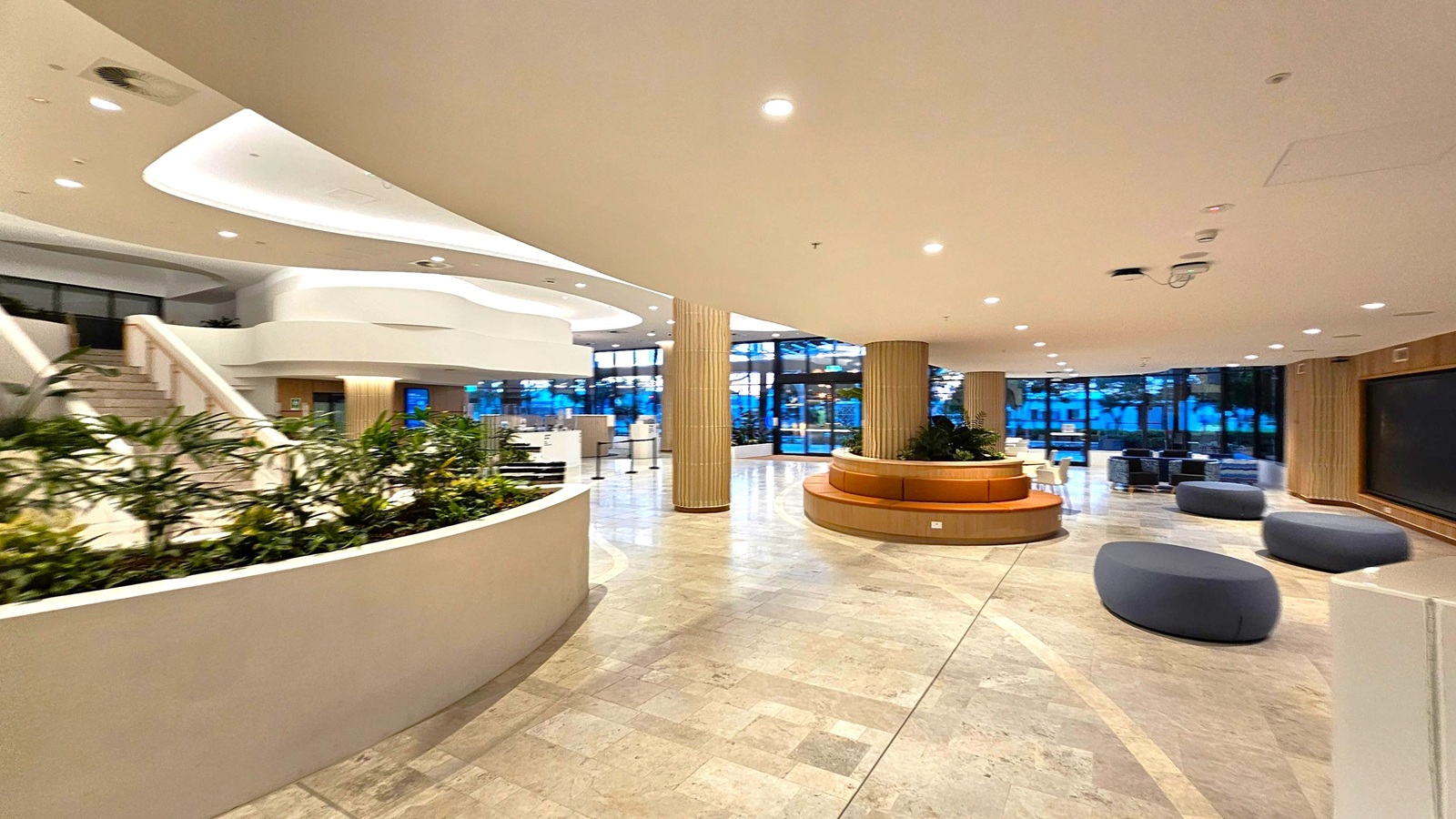 Spacious modern lobby with a light-coloured tiled floor. On the left, a curved planter with green plants and a staircase leading upwards. In the centre, a circular wooden seating area surrounds a column. On the right, several dark, oval-shaped seats are scattered. The background features large windows with a view of trees, and the ceiling is smooth with recessed lighting.