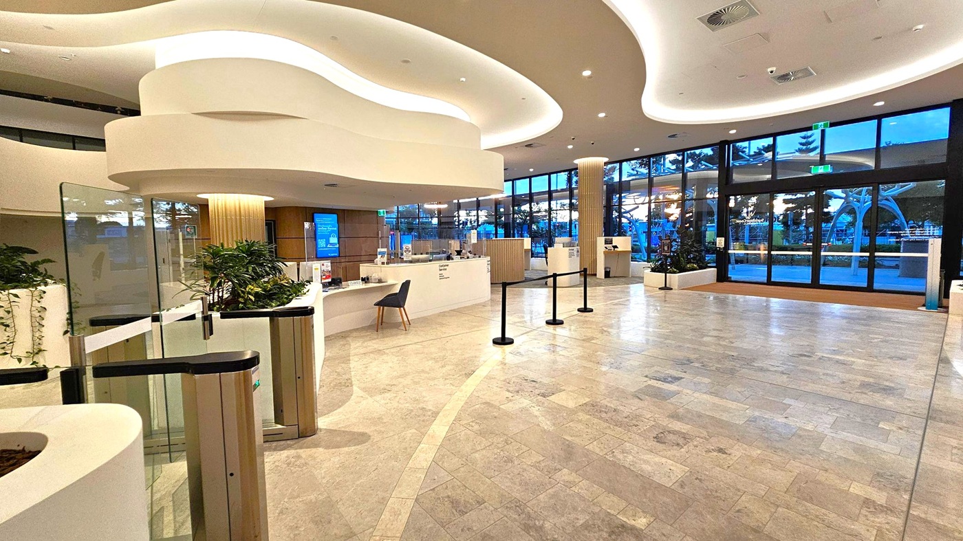 A modern interior with a spacious lobby area. On the left, there are curved white structures and a glass barrier with a gate. A small desk with a chair is positioned near the centre, surrounded by plants. The floor is tiled in a light colour with subtle patterns. On the right, large glass doors and windows provide a view of the outside, with trees visible. The ceiling features wavy, illuminated designs, and there are several overhead lights.