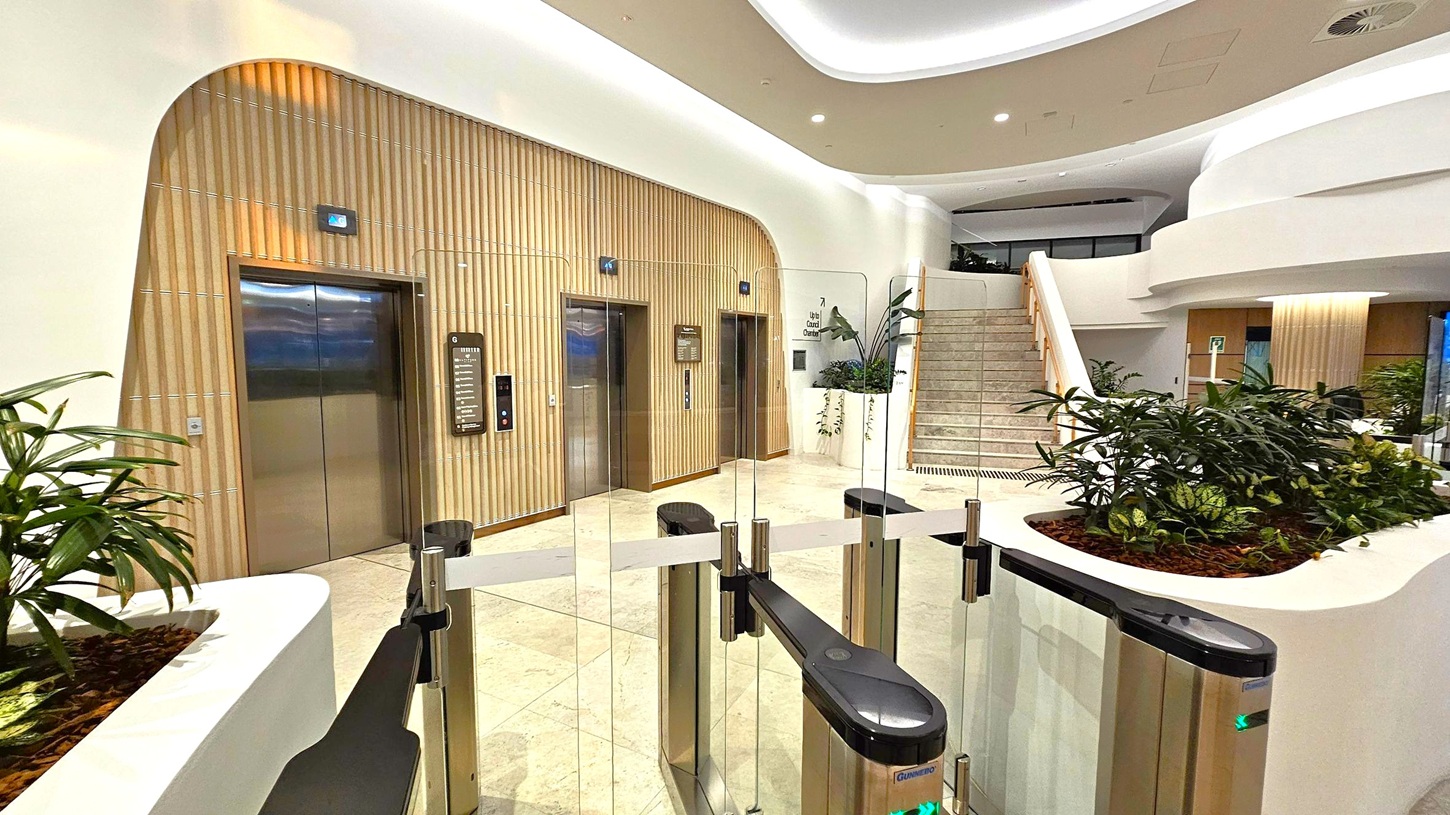 A modern interior with two lifts on the left, set in a wall with vertical wooden panelling. In the centre, a glass barrier with turnstiles. On the right, a staircase with a wooden handrail leads upwards. The foreground features planters with green plants, and the ceiling is smooth with recessed lighting. The floor is light-coloured and polished.
