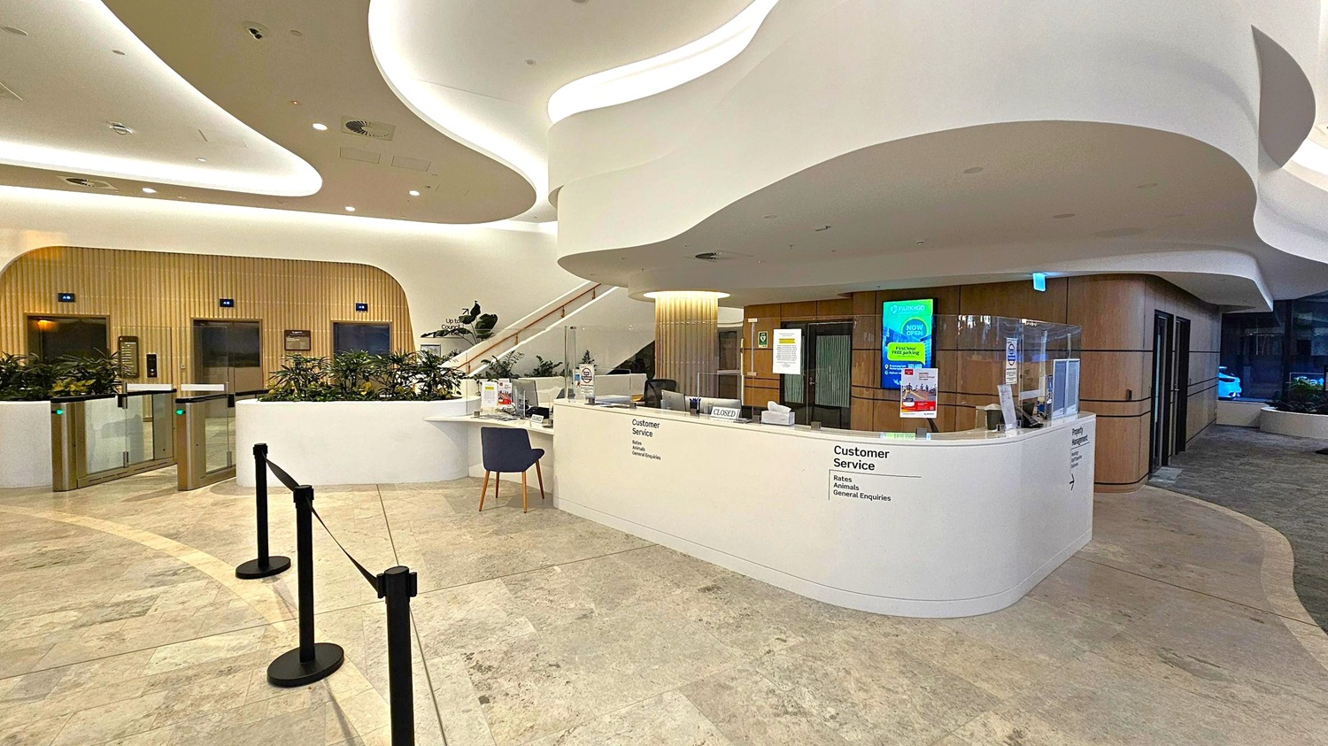 A modern interior with a customer service desk on the right, featuring a white counter and a black chair. Behind the desk, there are digital screens and informational posters. On the left, there are elevators with wooden panelling and a row of plants. The ceiling has a wavy design with recessed lighting. The floor is tiled in a light stone pattern, and there are black stanchions with ropes in the foreground.
