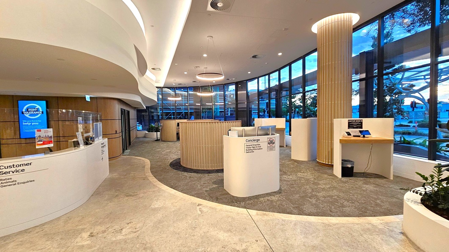 A modern interior space with a curved ceiling and recessed lighting. On the left, a customer service desk with a protective screen and digital signage. In the centre, a circular wooden structure and a concierge desk with brochures. On the right, a visitor sign-in station with tablets. Large floor-to-ceiling windows line the right side, offering a view of the outside. The floor is a mix of light and dark tiles, and a large column is present near the windows.