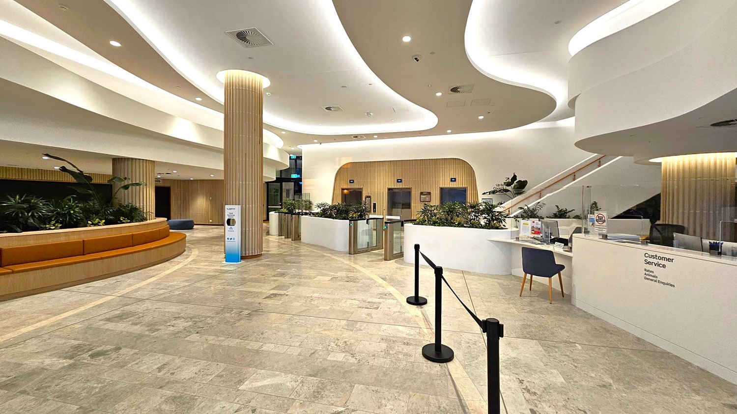 Spacious modern lobby with a curved design. On the left, a semi-circular brown bench is surrounded by plants. In the centre, a tall, ribbed column stands near a hand sanitiser station. The right side features a white customer service desk with a black chair. Behind the desk, a staircase ascends, and there are several plants and a barrier. The ceiling has flowing, wave-like patterns with recessed lighting, and the floor is tiled in a light stone pattern.