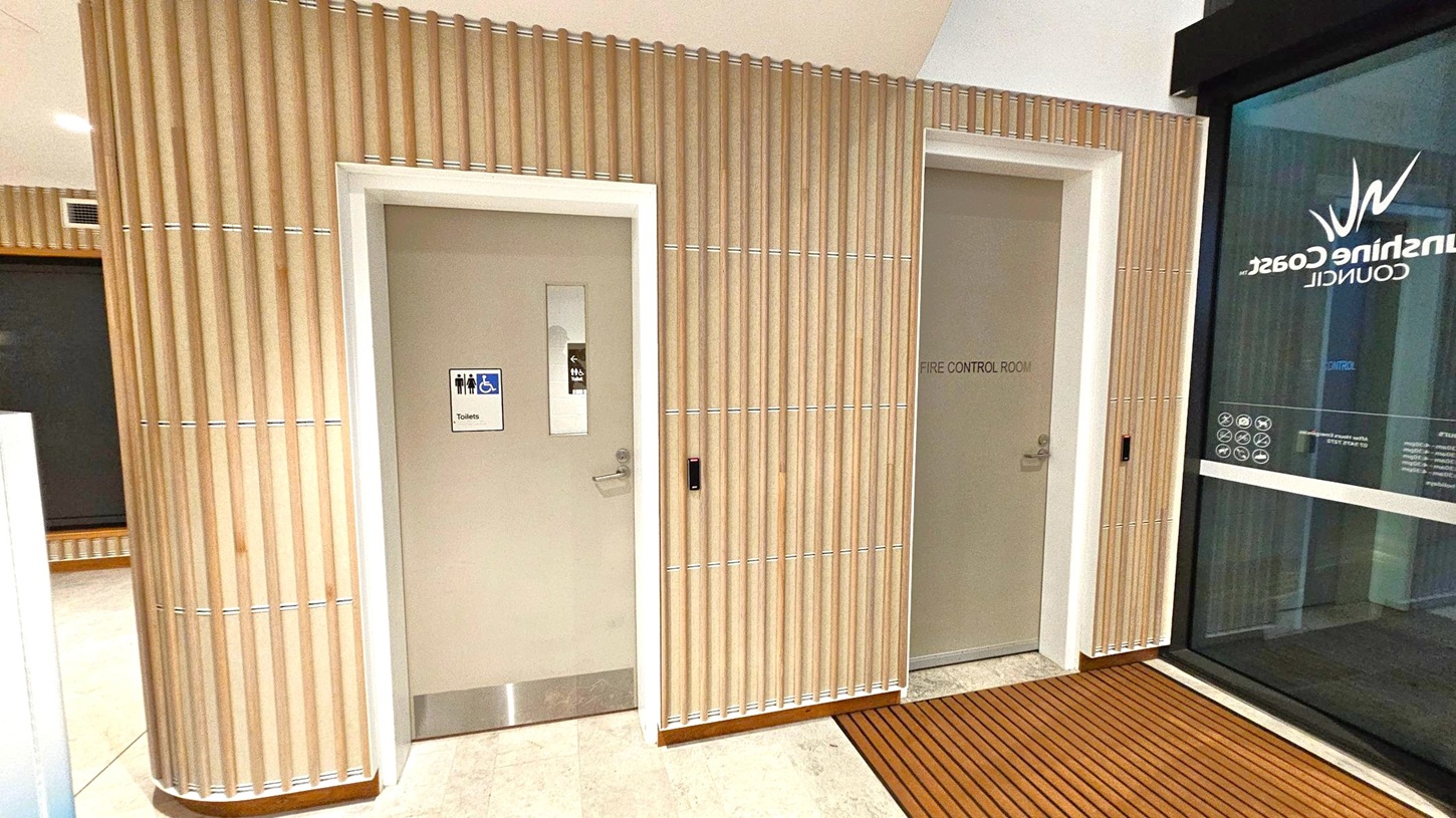 A hallway with two doors on a wall featuring vertical wooden slats. The left door has a sign indicating toilets, including accessible facilities, and a small window. The right door is labelled "Fire Control Room." To the right, a glass panel displays the words "Sunshine Coast Council" and various symbols. The floor is tiled, transitioning to a wooden slat mat near the glass panel. The ceiling is plain with recessed lighting.