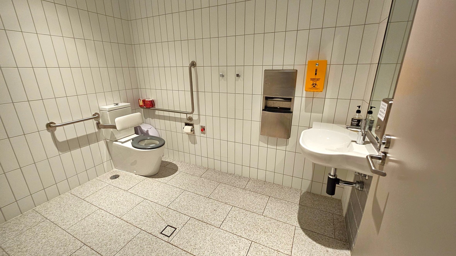 A public accessible toilet with white tiled walls and a speckled grey floor. On the left, a toilet with a blue seat is equipped with support rails. Above it, a red emergency pull cord and a toilet roll holder are visible. In the centre, a stainless steel dispenser is mounted on the wall. On the right, a white sink with a mirror above it and soap dispensers is present. A yellow sign is affixed to the wall near the sink.