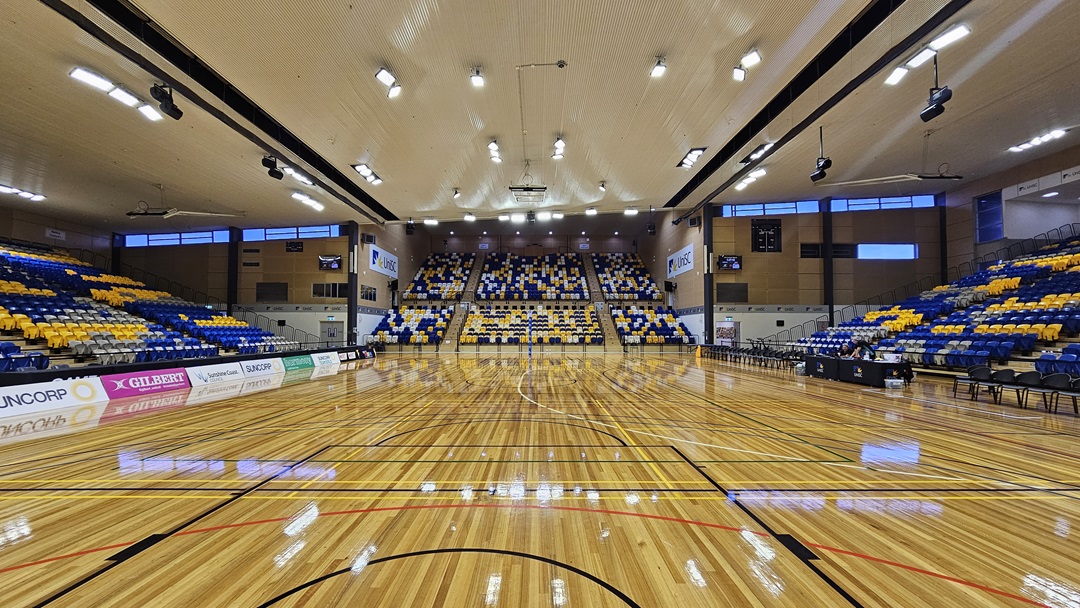 On the left, there are rows of blue and yellow seats with advertising banners along the lower section, including brands like HCF and Suncorp. The middle features a large seating area with blue, yellow, and white seats arranged to spell "USC" and "SPARTANS." The right side has more seating in the same colours, with additional advertising banners. The foreground shows a polished wooden basketball court with green and yellow lines. The ceiling is high with recessed lighting and ventilation ducts.