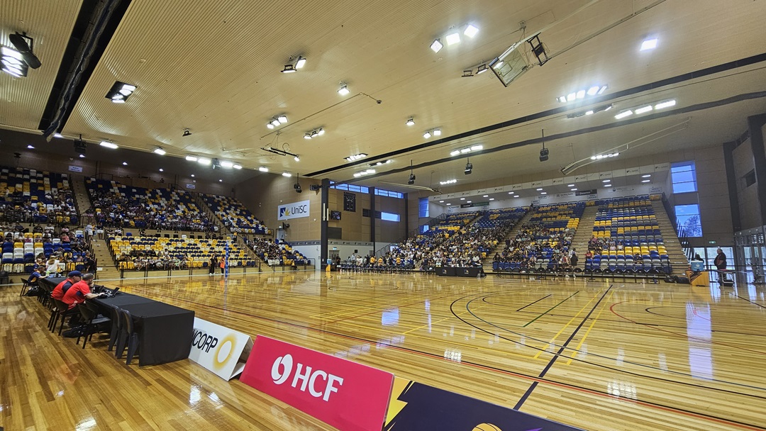 On the left, there are rows of blue, yellow, and white seats with advertising banners for Suncorp and HCF along the lower section. A table with people seated is positioned near the court. The middle features a polished wooden basketball court with lines and a central logo. The right side has more seating in blue, yellow, and white, with additional advertising banners. The ceiling is high with recessed lighting and ventilation ducts.