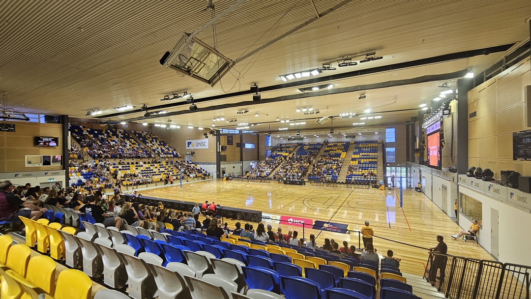 On the left, there are rows of blue, yellow, and white seats with spectators. The middle features a polished wooden basketball court with lines and a central logo. The right side has more seating in blue, yellow, and white, with additional spectators and advertising banners. The background includes more seating, large windows, and a scoreboard. The ceiling is high with recessed lighting, ventilation ducts, and a large suspended scoreboard above the court.