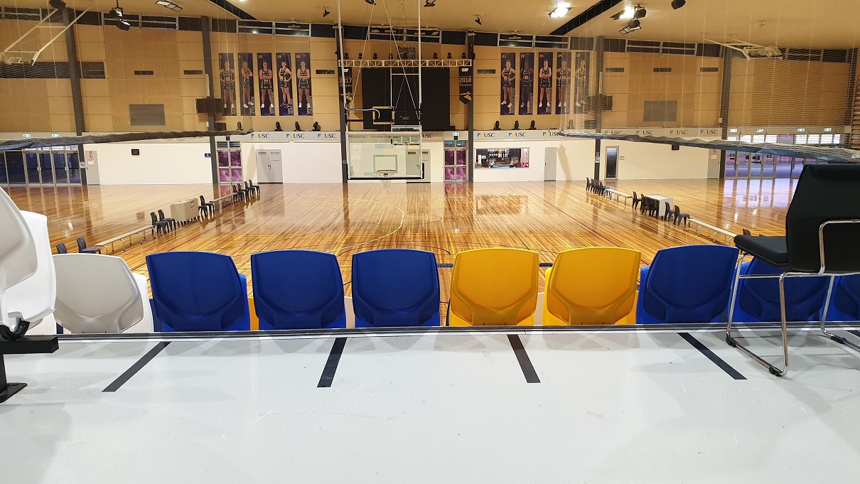 On the left, there are white and blue seats. The middle features a row of blue and yellow seats overlooking a polished wooden basketball court with black lines. The right side has more blue and black seats. The background includes the basketball court, with chairs and tables along the sides, large banners on the far wall, and a scoreboard. The ceiling is high with recessed lighting, ventilation ducts, and suspended basketball hoops. The foreground shows a white floor with black markings.