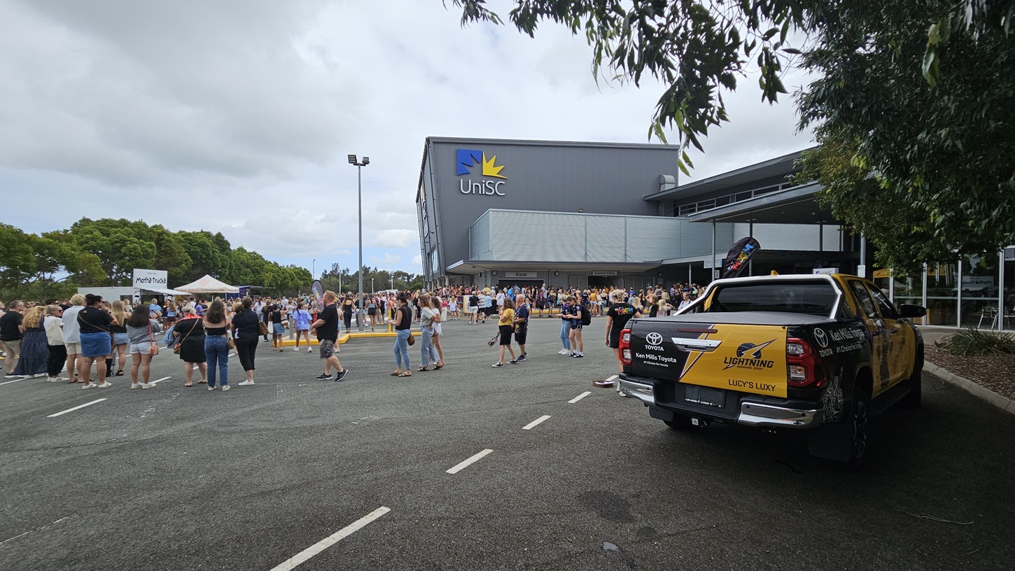 On the left, there is a crowd of people gathered in an open area with a few tents and trees in the background. In the middle, more people are standing and walking near the entrance of a large building with a "UniSC" sign and a yellow star logo. The right side features a yellow and black utility vehicle with "Sunshine Coast Lightning" branding parked on an asphalt surface. The background includes the building's facade and more trees.