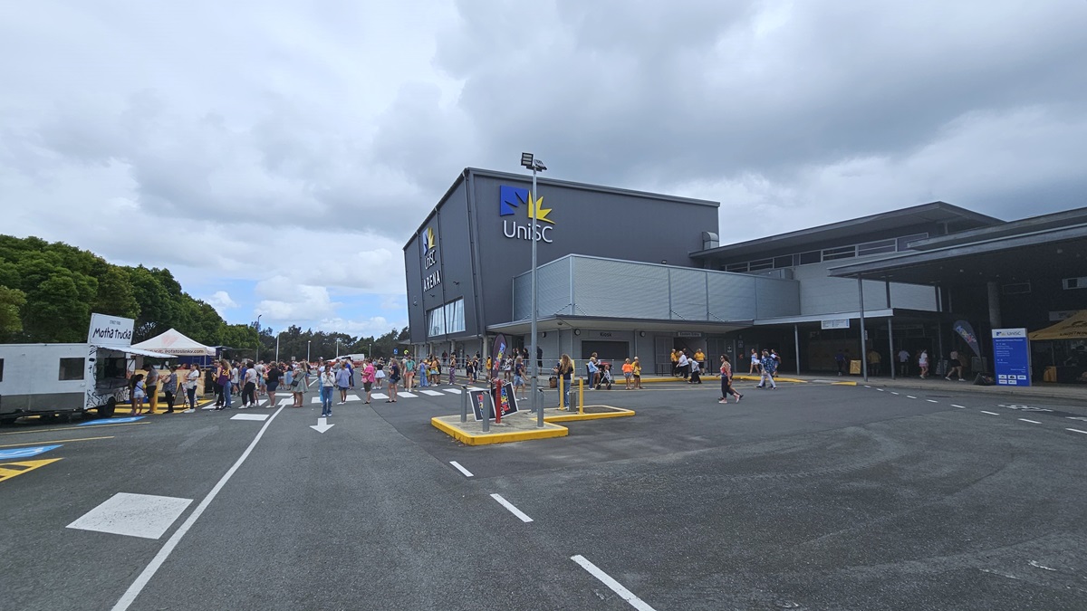 On the left, a white food truck with an open serving window is surrounded by people, next to a white tent where more individuals gather. In the center, a large grey building sports a "UniSC" sign and a yellow star logo, with people walking towards the entrance and others nearby. The right side displays a covered walkway with people entering and exiting the building. The foreground features an asphalt car park with white-lined parking spaces and yellow kerbs around pedestrian zones.